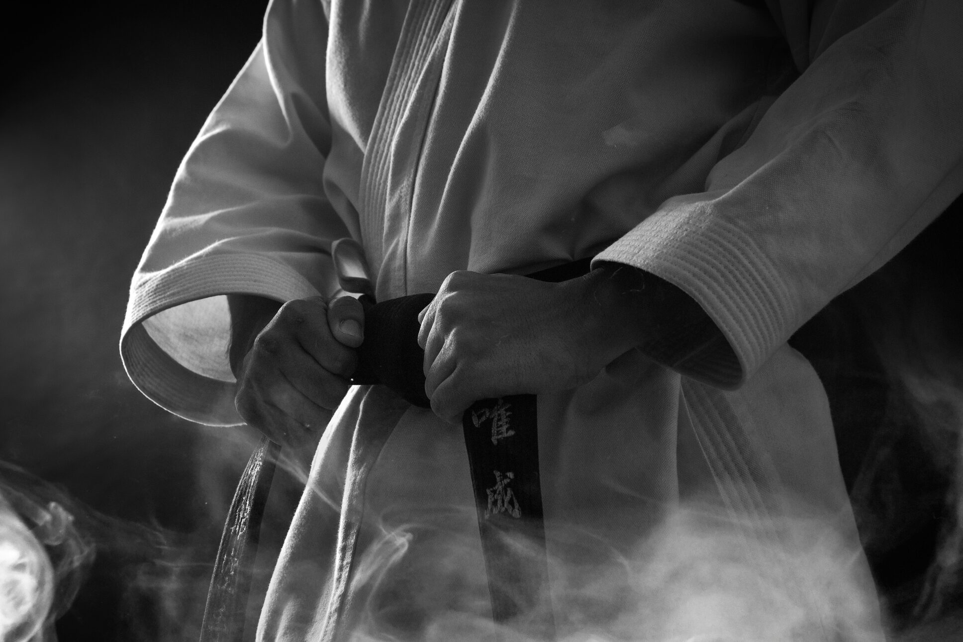 Gros plan en noir et blanc sur un karatéka en kimono blanc nouant sa ceinture noire brodée de kanji.